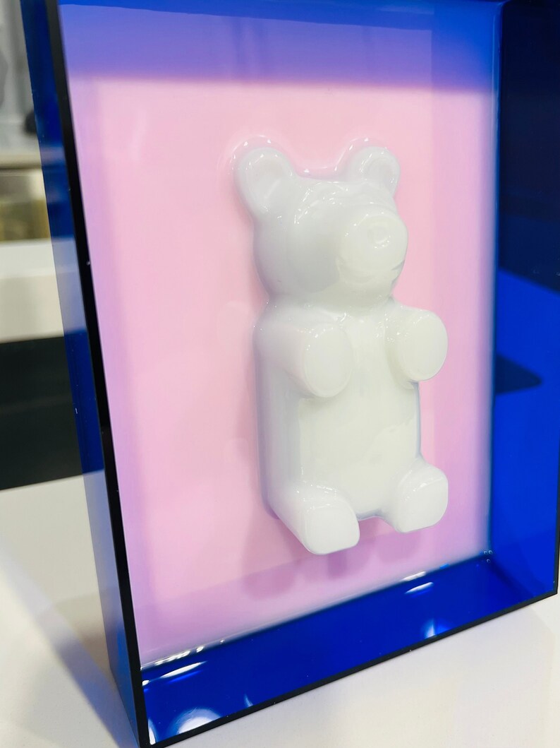 Puede incluir: Una figura tridimensional de oso de goma blanco montada sobre un fondo rosa plano, encerrada dentro de un marco rectangular azul transl&uacute;cido profundo. El oso tiene un acabado suave y brillante y est&aacute; centrado sobre el fondo rosa claro.