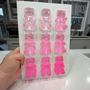 Pu&ograve; includere: Uno stampo rettangolare bianco pieno di nove orsetti gommosi rosa. Gli orsetti gommosi sono disposti in tre file da tre, con diverse tonalit&agrave; di rosa. Lo stampo &egrave; tenuto in mano e lo sfondo &egrave; una cucina.