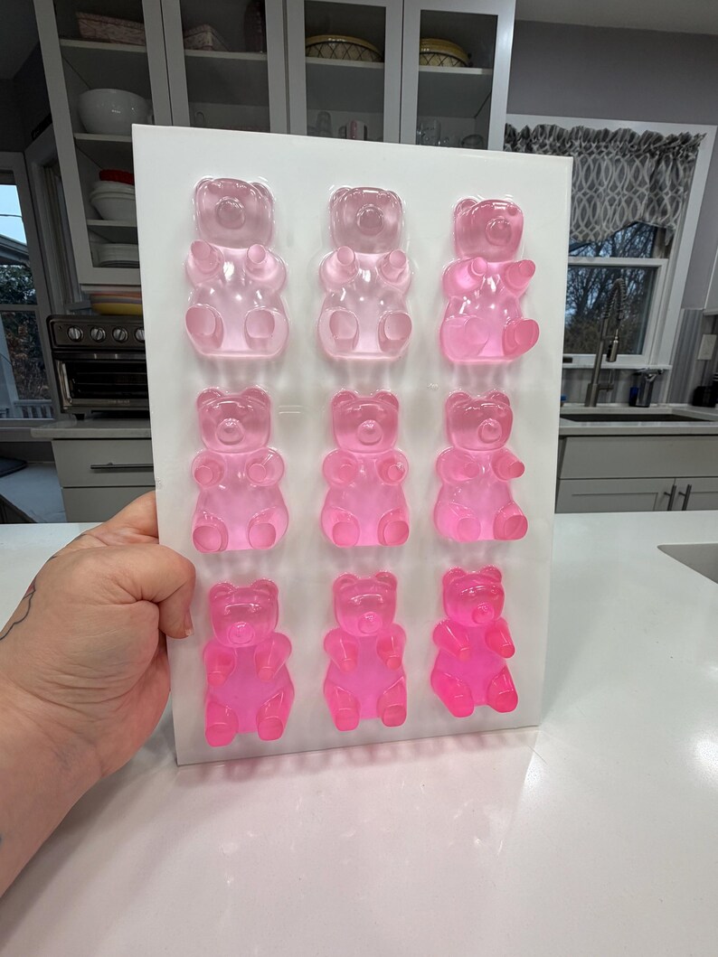 Pu&ograve; includere: Uno stampo rettangolare bianco pieno di nove orsetti gommosi rosa. Gli orsetti gommosi sono disposti in tre file da tre, con diverse tonalit&agrave; di rosa. Lo stampo &egrave; tenuto in mano e lo sfondo &egrave; una cucina.