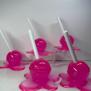 Puede incluir: Varias piruletas rosas con palitos blancos, que parecen derretirse sobre una superficie blanca. Las piruletas tienen un acabado brillante y est&aacute;n dispuestas en grupo, creando una exhibici&oacute;n l&uacute;dica y colorida.