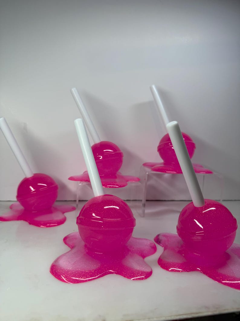Puede incluir: Varias piruletas rosas con palitos blancos, que parecen derretirse sobre una superficie blanca. Las piruletas tienen un acabado brillante y est&aacute;n dispuestas en grupo, creando una exhibici&oacute;n l&uacute;dica y colorida.