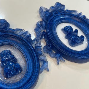 Op de afbeelding: Twee blauwe glitter fotolijstjes met witte binnenkant en een blauwe glitter berenfiguur in elk lijstje. De lijstjes zijn versierd met wit lint.