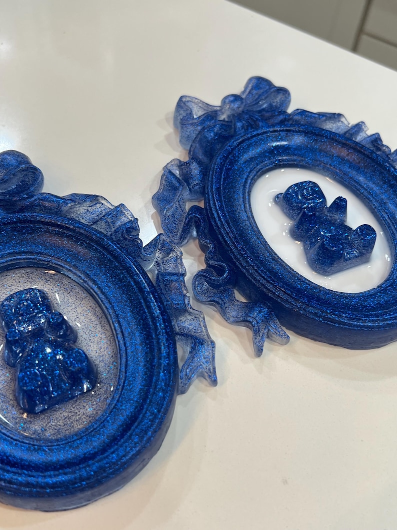 Op de afbeelding: Twee blauwe glitter fotolijstjes met witte binnenkant en een blauwe glitter berenfiguur in elk lijstje. De lijstjes zijn versierd met wit lint.
