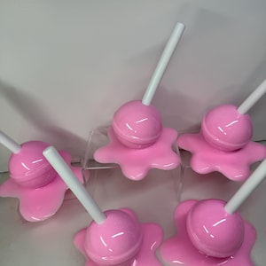 Op de afbeelding: Een verzameling van zes roze objecten in de vorm van lolly's. Elke lolly heeft een ronde, roze snoepkop en een witte stok. Het snoepgoed rust op een roze, gesmolten uitziende basis. De objecten zijn gerangschikt op een helder, verhoogd oppervlak.