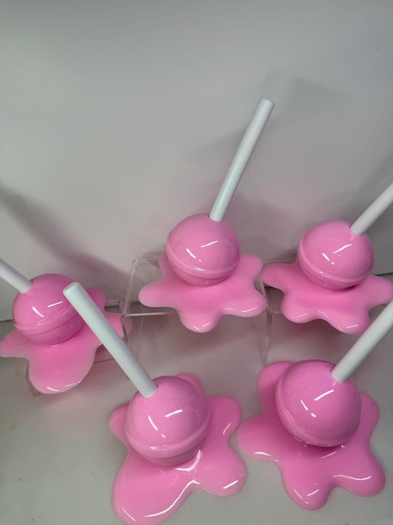 Op de afbeelding: Een verzameling van zes roze objecten in de vorm van lolly's. Elke lolly heeft een ronde, roze snoepkop en een witte stok. Het snoepgoed rust op een roze, gesmolten uitziende basis. De objecten zijn gerangschikt op een helder, verhoogd oppervlak.