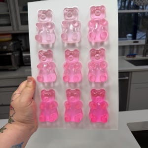Pu&ograve; includere: Uno stampo in silicone bianco riempito con nove caramelle gommose a forma di orsetto rosa. Gli orsetti gommosi sono disposti in tre file da tre, con diverse tonalit&agrave; di rosa. Lo stampo &egrave; tenuto da una mano.