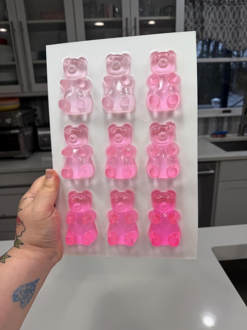 Pu&ograve; includere: Uno stampo in silicone bianco riempito con nove caramelle gommose a forma di orsetto rosa. Gli orsetti gommosi sono disposti in tre file da tre, con diverse tonalit&agrave; di rosa. Lo stampo &egrave; tenuto da una mano.
