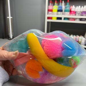 May include: A clear plastic heart-shaped container filled with colorful slime, including pink, blue, yellow, and orange. The slime is surrounded by a clear liquid.
