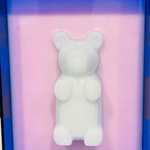 Puede incluir: Una figura de oso de goma blanca y transl&uacute;cida en tres dimensiones, centrada sobre un fondo rectangular rosa claro, todo ello encerrado en un marco azul profundo que crea un contraste audaz con los tonos pastel suaves del oso central.