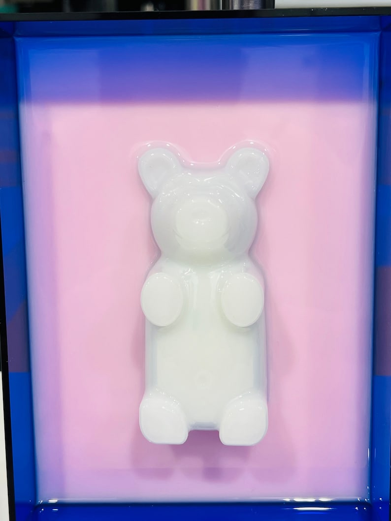 Puede incluir: Una figura de oso de goma blanca y transl&uacute;cida en tres dimensiones, centrada sobre un fondo rectangular rosa claro, todo ello encerrado en un marco azul profundo que crea un contraste audaz con los tonos pastel suaves del oso central.