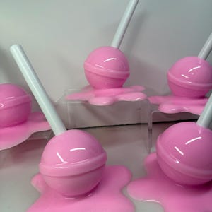 Op de afbeelding: Roze lolly's met witte stokjes, die lijken te smelten op een oppervlak. De lolly's hebben een ronde vorm met een band in het midden. Het gesmolten snoepgoed is een lichtere tint roze.