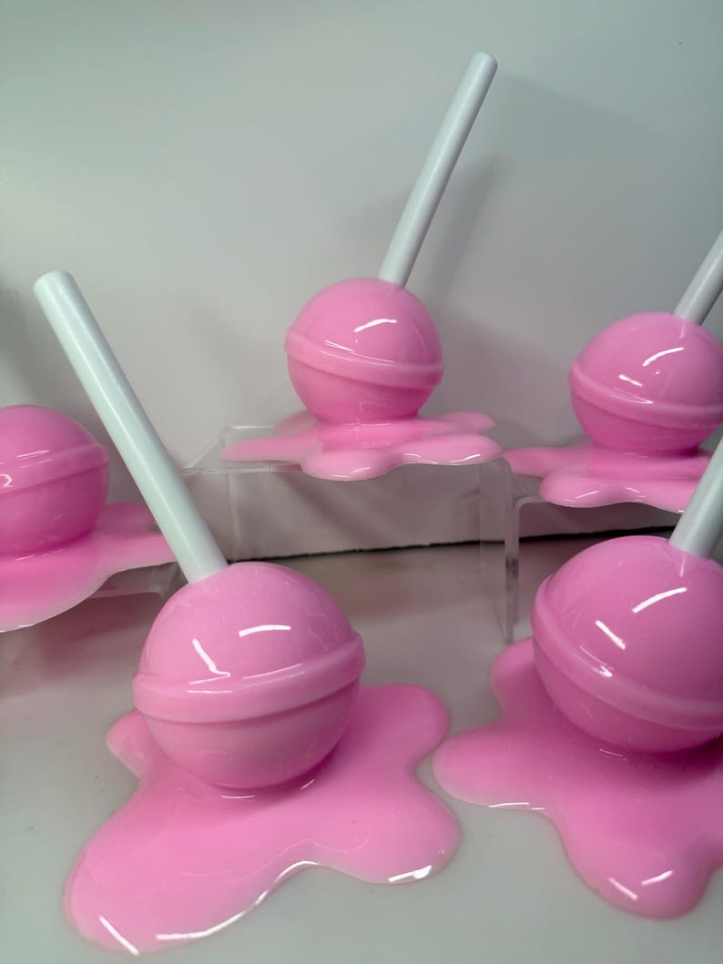Op de afbeelding: Roze lolly's met witte stokjes, die lijken te smelten op een oppervlak. De lolly's hebben een ronde vorm met een band in het midden. Het gesmolten snoepgoed is een lichtere tint roze.