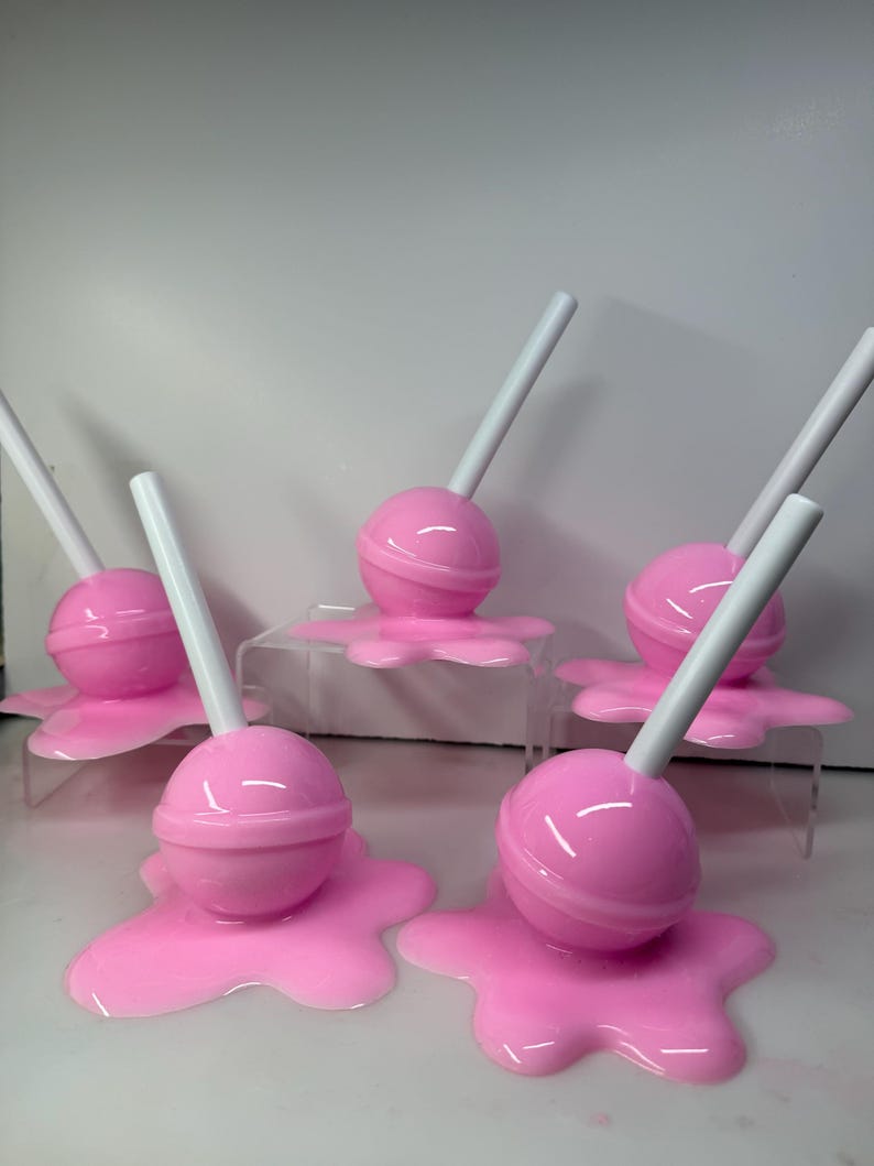 Op de afbeelding: Roze lolly's met witte stokjes, die lijken te smelten op een bijpassende roze basis. De lolly's zijn geplaatst op heldere, verhoogde platforms. De afbeelding toont een speels, snoep-thema ontwerp.