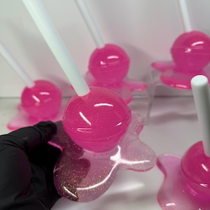 Puede incluir: Varias piezas de arte de resina en forma de piruletas rosas y brillantes con palitos blancos. Las piruletas tienen una parte superior redonda y brillante y una base con aspecto de derretida. La base tiene un efecto brillante. La imagen est&aacute; tomada de cerca.