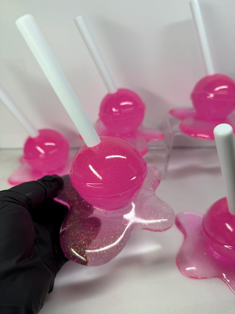 Puede incluir: Varias piezas de arte de resina en forma de piruletas rosas y brillantes con palitos blancos. Las piruletas tienen una parte superior redonda y brillante y una base con aspecto de derretida. La base tiene un efecto brillante. La imagen est&aacute; tomada de cerca.