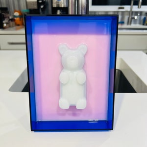 Puede incluir: Un marco rectangular con un borde azul profundo y un panel interior rosa claro. Centrada en el panel hay una figura tridimensional de osito de goma blanco opaco que sobresale de la superficie, creando un efecto visual de capas.
