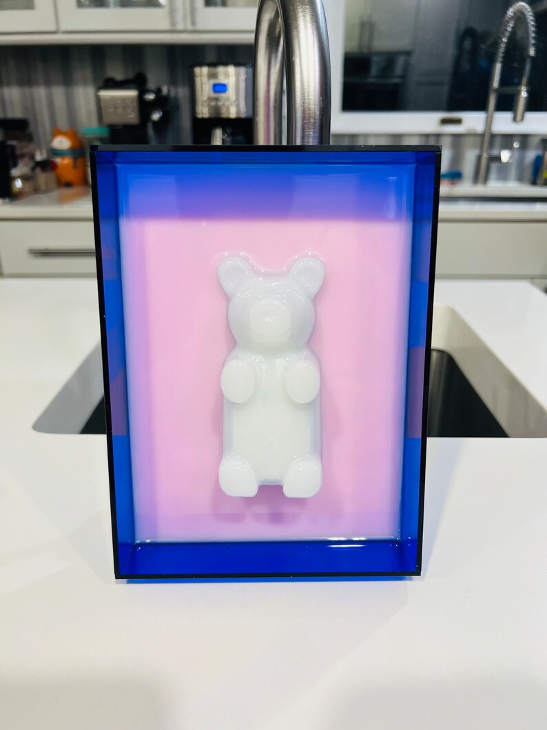 Puede incluir: Un marco rectangular con un borde azul profundo y un panel interior rosa claro. Centrada en el panel hay una figura tridimensional de osito de goma blanco opaco que sobresale de la superficie, creando un efecto visual de capas.