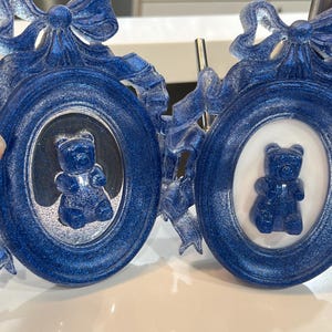 Op de afbeelding: Twee blauwe glitter fotolijstjes met sierlijke ontwerpen en witte achtergronden. Elk lijstje heeft een blauwe gummybeer erin.