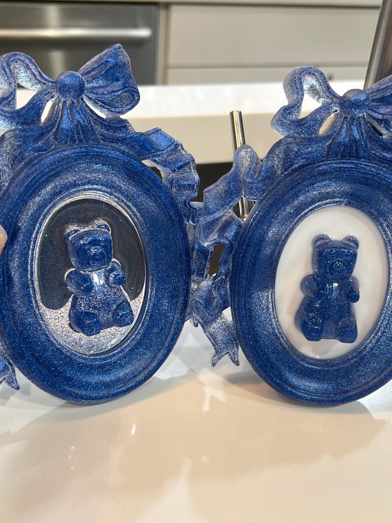 Op de afbeelding: Twee blauwe glitter fotolijstjes met sierlijke ontwerpen en witte achtergronden. Elk lijstje heeft een blauwe gummybeer erin.
