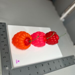 May include: Three colorful, fruit-shaped candles on a white surface. The candles are shaped like raspberries and cherries. The first candle is orange, the second is pink, and the third is red. Each candle has a green stem. A ruler is placed next to the candles.