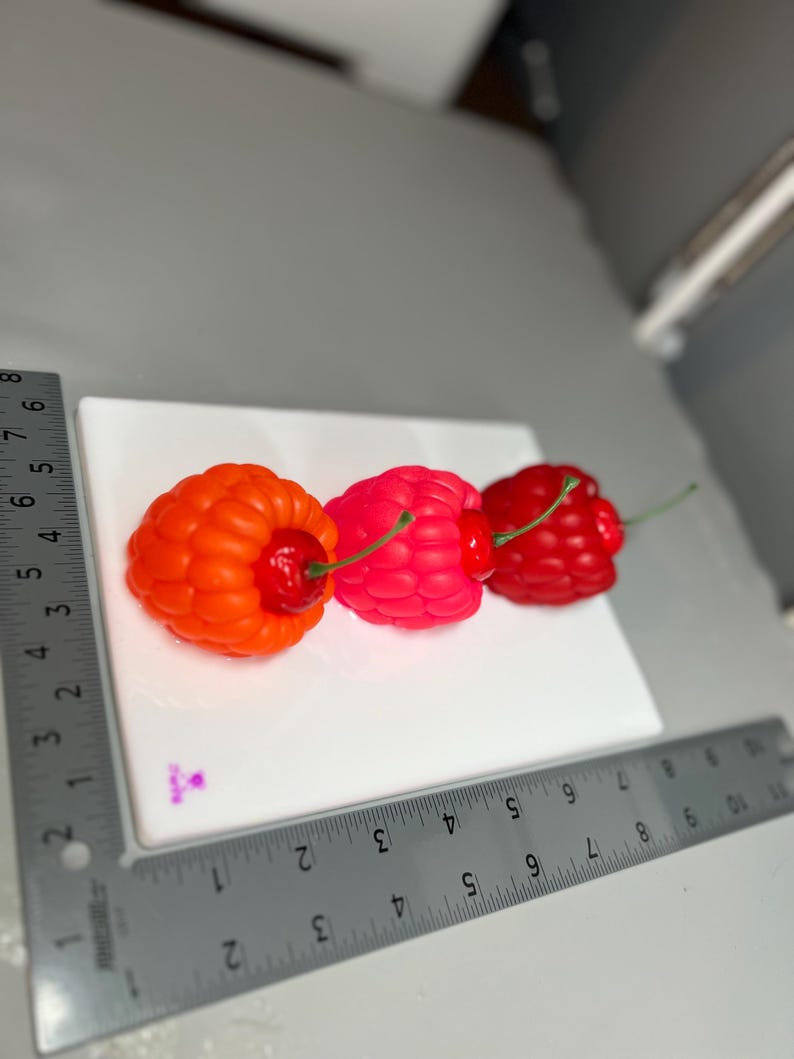 May include: Three colorful, fruit-shaped candles on a white surface. The candles are shaped like raspberries and cherries. The first candle is orange, the second is pink, and the third is red. Each candle has a green stem. A ruler is placed next to the candles.