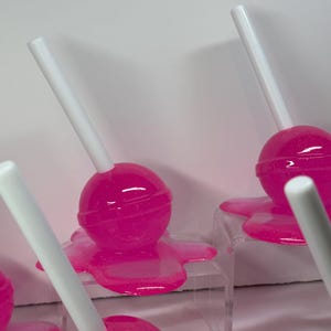 Puede incluir: Se muestran varias piruletas de color rosa brillante con palitos blancos. Las piruletas parecen estar derriti&eacute;ndose sobre peque&ntilde;os expositores transparentes. El fondo es una superficie blanca lisa.