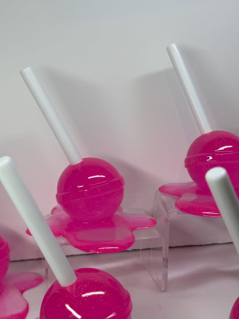 Puede incluir: Se muestran varias piruletas de color rosa brillante con palitos blancos. Las piruletas parecen estar derriti&eacute;ndose sobre peque&ntilde;os expositores transparentes. El fondo es una superficie blanca lisa.