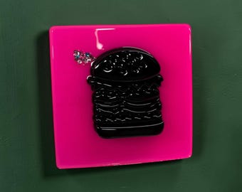 May include: A square, bright pink wall plaque featuring a glossy, raised black burger icon in the center. A small cluster of iridescent, multi-colored sequins is attached to the upper left side of the burger, adding a textured, reflective detail.