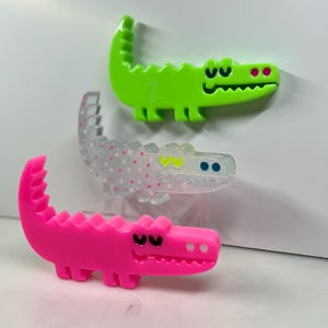 May include: Three colorful alligator-shaped pins. The top pin is bright green, the middle one is clear with pink dots, and the bottom one is hot pink. Each pin has a smiling face and a textured back.