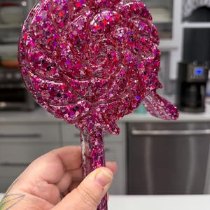 May include: A lollipop-shaped object featuring a swirling, circular head and a straight handle, entirely covered in dense, multi-sized pink and magenta glitter. The surface is glossy and smooth, reflecting light across the textured, sparkling finish.