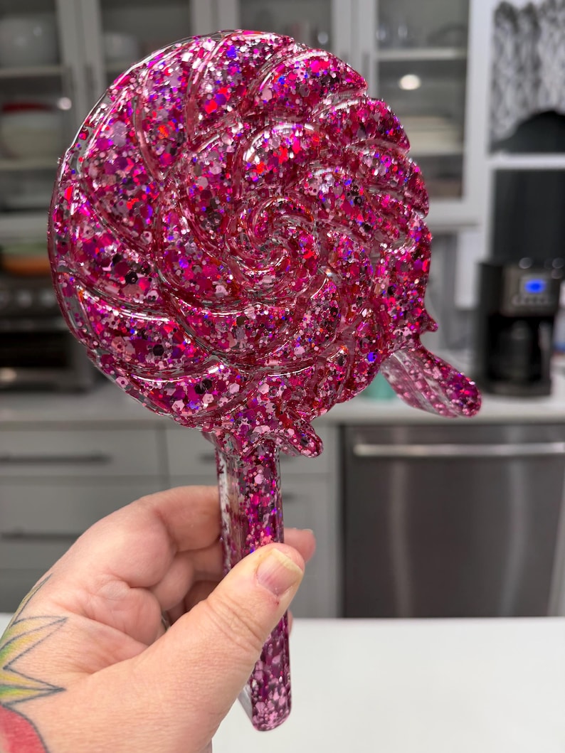 May include: A lollipop-shaped object featuring a swirling, circular head and a straight handle, entirely covered in dense, multi-sized pink and magenta glitter. The surface is glossy and smooth, reflecting light across the textured, sparkling finish.