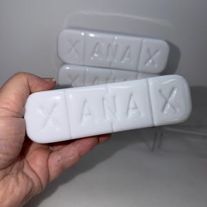 以下が含まれることがあります： 表面に「XANA」の文字がエンボス加工された、白い長方形の錠剤型の物体。物体は積み重ねられており、1つに焦点が当てられています。背景は白です。