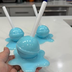 Könnte beinhalten: Drei hellblaue Objekte in Lutscherform mit weißen Stäben. Jeder Lutscher sitzt auf einer passenden hellblauen, geschmolzen aussehenden Basis. Die Objekte befinden sich auf einer weißen Oberfläche, mit einem verschwommenen Küchenhintergrund.