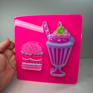 May include: A bright pink square with a burger and milkshake design. The burger has pink glitter and purple accents. The milkshake is purple and pink with a green cherry on top. The design is on a bright pink background.