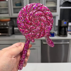 May include: A lollipop-shaped object made of translucent pink resin filled with dense, multi-sized pink and magenta glitter. The circular head features a swirling pattern, and a small drip of resin extends from the bottom edge near the handle.