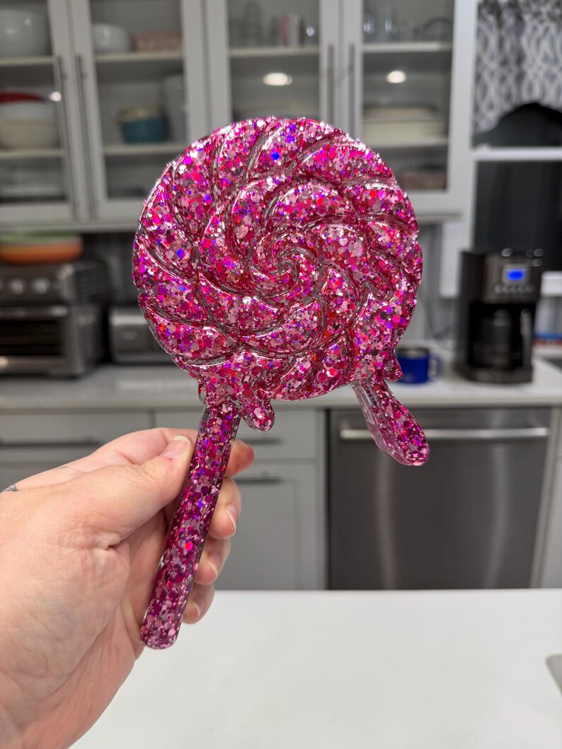 May include: A lollipop-shaped object made of translucent pink resin filled with dense, multi-sized pink and magenta glitter. The circular head features a swirling pattern, and a small drip of resin extends from the bottom edge near the handle.
