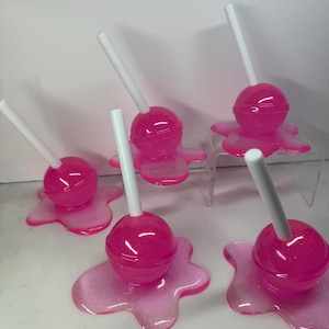 Puede incluir: Se muestran varias piruletas rosas con palitos blancos. Cada piruleta se asienta sobre una base rosa brillante con aspecto derretido. Las piruletas son de un color rosa vibrante y las bases tienen un efecto brillante.