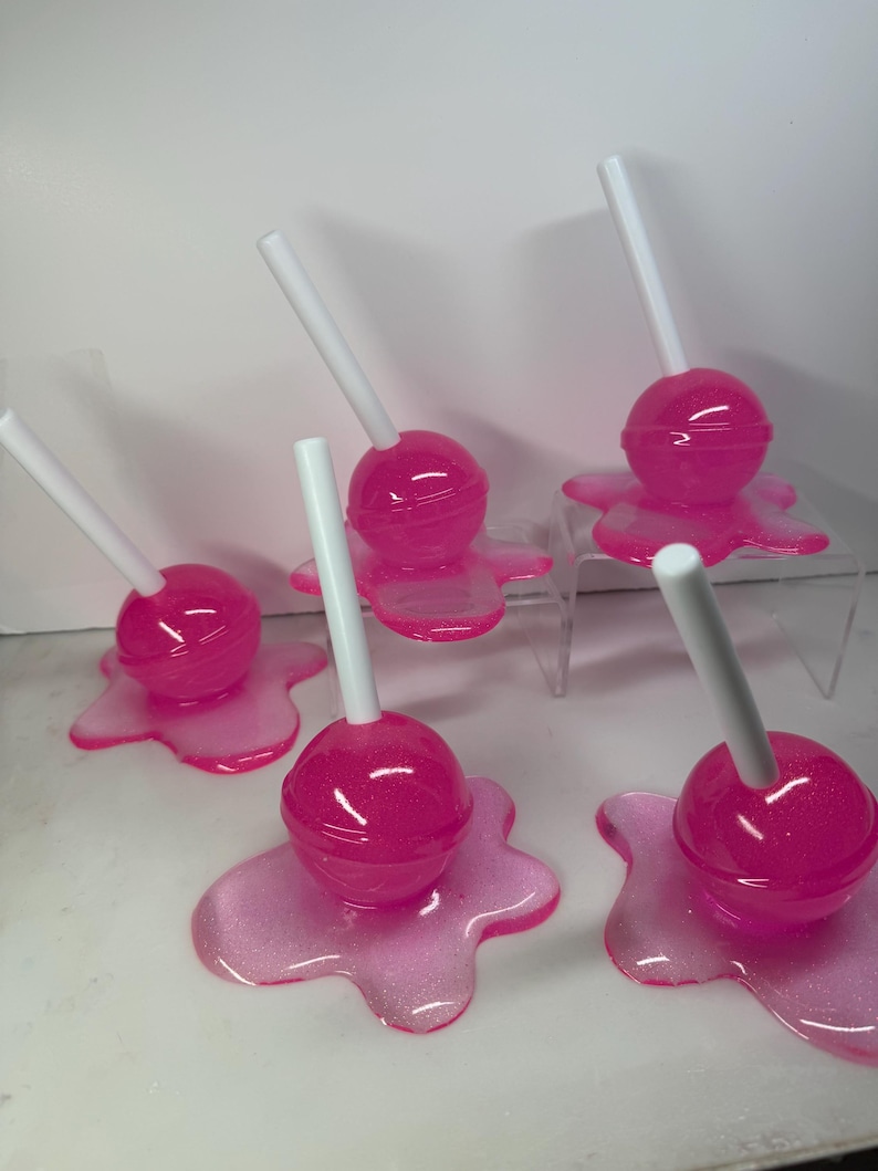 Puede incluir: Se muestran varias piruletas rosas con palitos blancos. Cada piruleta se asienta sobre una base rosa brillante con aspecto derretido. Las piruletas son de un color rosa vibrante y las bases tienen un efecto brillante.