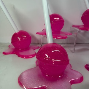 Puede incluir: Varias piruletas de color rosa brillante con palitos blancos, cada una sobre una base rosa brillante y derretida a juego. Las piruletas est&aacute;n dispuestas sobre una superficie blanca, creando una presentaci&oacute;n divertida. El caramelo es de un rosa vibrante y brillante.