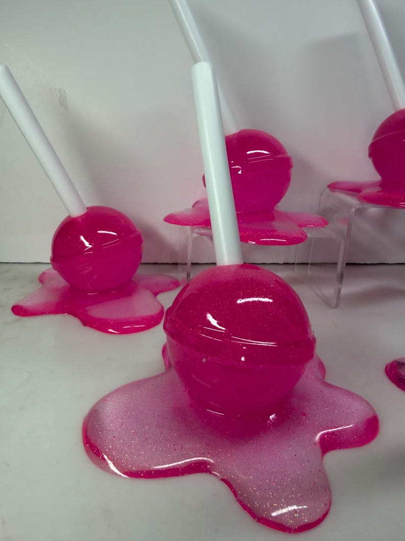 Puede incluir: Varias piruletas de color rosa brillante con palitos blancos, cada una sobre una base rosa brillante y derretida a juego. Las piruletas est&aacute;n dispuestas sobre una superficie blanca, creando una presentaci&oacute;n divertida. El caramelo es de un rosa vibrante y brillante.