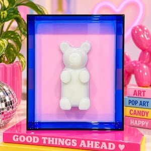 Puede incluir: Una pieza de arte mural rectangular con un marco de acr&iacute;lico azul transl&uacute;cido que rodea un fondo rosa claro. Una figura de oso de goma blanca en tres dimensiones est&aacute; centrada sobre la superficie rosa, creando una exhibici&oacute;n decorativa moderna.