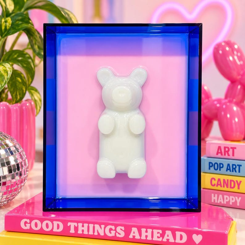 Puede incluir: Una pieza de arte mural rectangular con un marco de acr&iacute;lico azul transl&uacute;cido que rodea un fondo rosa claro. Una figura de oso de goma blanca en tres dimensiones est&aacute; centrada sobre la superficie rosa, creando una exhibici&oacute;n decorativa moderna.