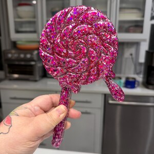 May include: A handheld mirror shaped like a swirling lollipop, crafted from translucent pink resin embedded with dense, multi-sized holographic glitter. The circular head features a spiral design with a small, dripping accent, attached to a straight handle.