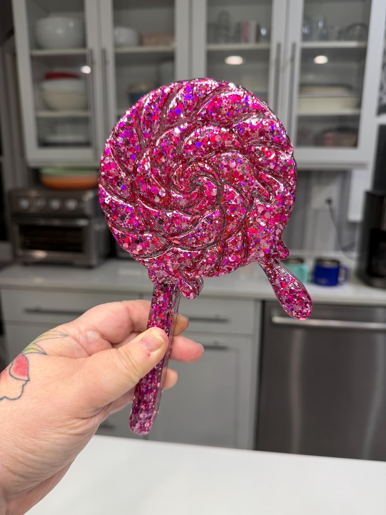 May include: A handheld mirror shaped like a swirling lollipop, crafted from translucent pink resin embedded with dense, multi-sized holographic glitter. The circular head features a spiral design with a small, dripping accent, attached to a straight handle.