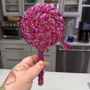 May include: A circular lollipop-shaped object featuring a swirling pattern filled with vibrant pink glitter and holographic sequins. The piece includes a straight handle extending from the base, all crafted with a glossy, resin-like finish.