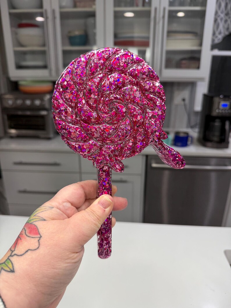 May include: A circular lollipop-shaped object featuring a swirling pattern filled with vibrant pink glitter and holographic sequins. The piece includes a straight handle extending from the base, all crafted with a glossy, resin-like finish.