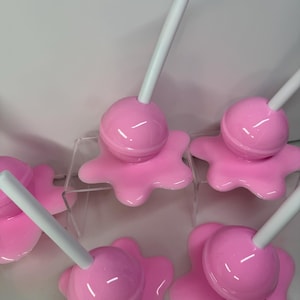 Op de afbeelding: Roze lolly-vormige decoratieve objecten met witte stokjes. De glanzende lolly's staan op een bijpassende, gesmolten roze basis. De afbeelding toont meerdere van deze items, wat wijst op een set of verzameling.