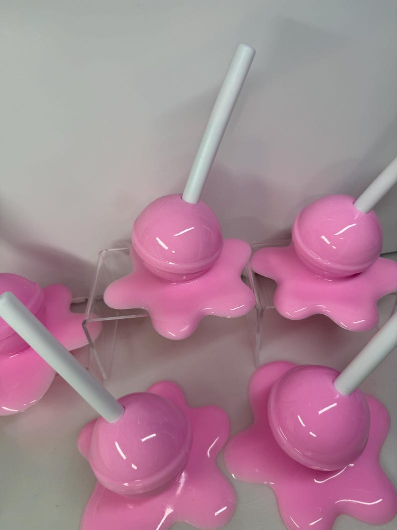 Op de afbeelding: Roze lolly-vormige decoratieve objecten met witte stokjes. De glanzende lolly's staan op een bijpassende, gesmolten roze basis. De afbeelding toont meerdere van deze items, wat wijst op een set of verzameling.
