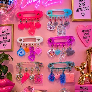 May include: Six large safety pin wall displays in pink, white, blue, and glitter finishes. Each pin features hanging charms including hearts, keys, and birdcages. The display is set against a pink wall with neon text reading Candy Cabinet.