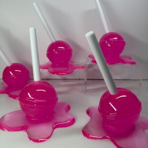 Puede incluir: Varias piruletas de color rosa brillante con palitos blancos. Cada piruleta se asienta sobre una base rosa con aspecto de derretida. Las piruletas tienen un aspecto brillante y las bases tienen un efecto brillante.